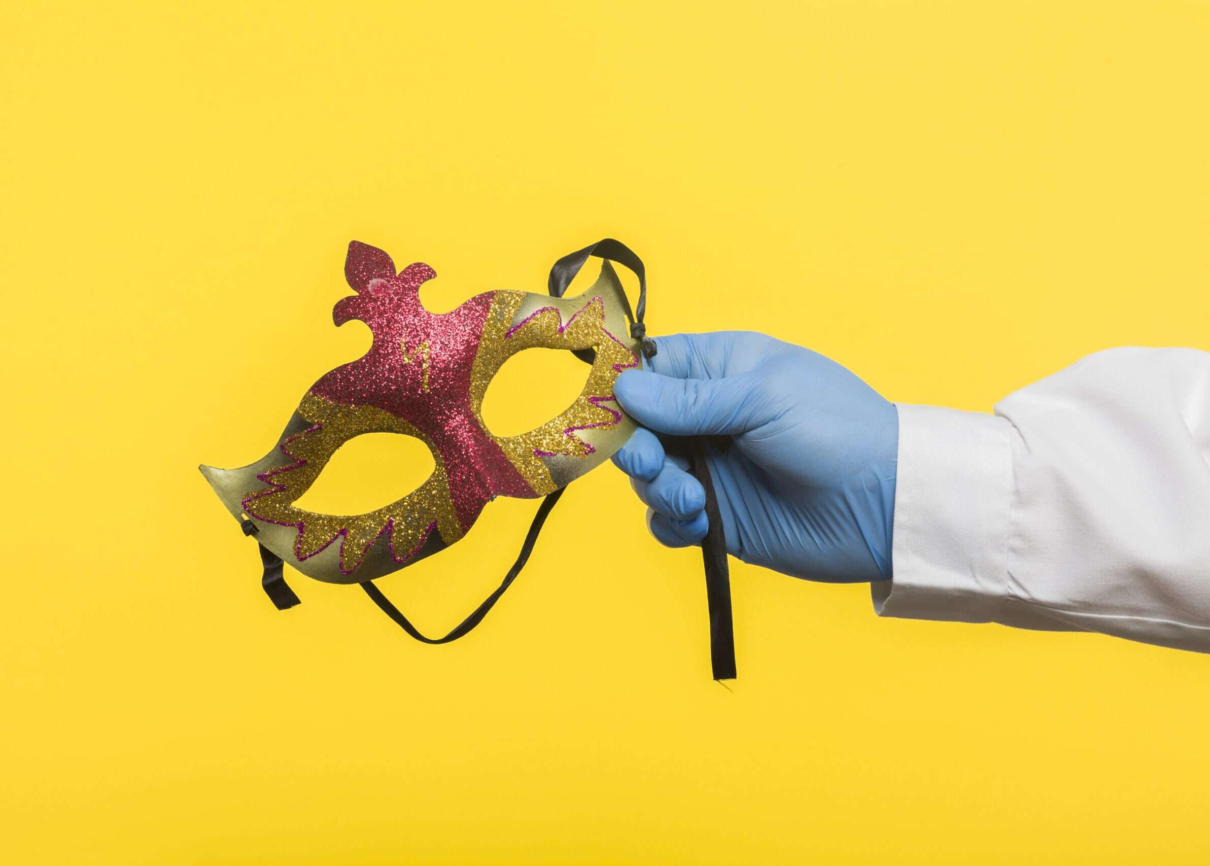 Closeup side view profile of a human hand in blue surgical gloves, holding a carnival mask model on the face, isolated on a yellow background